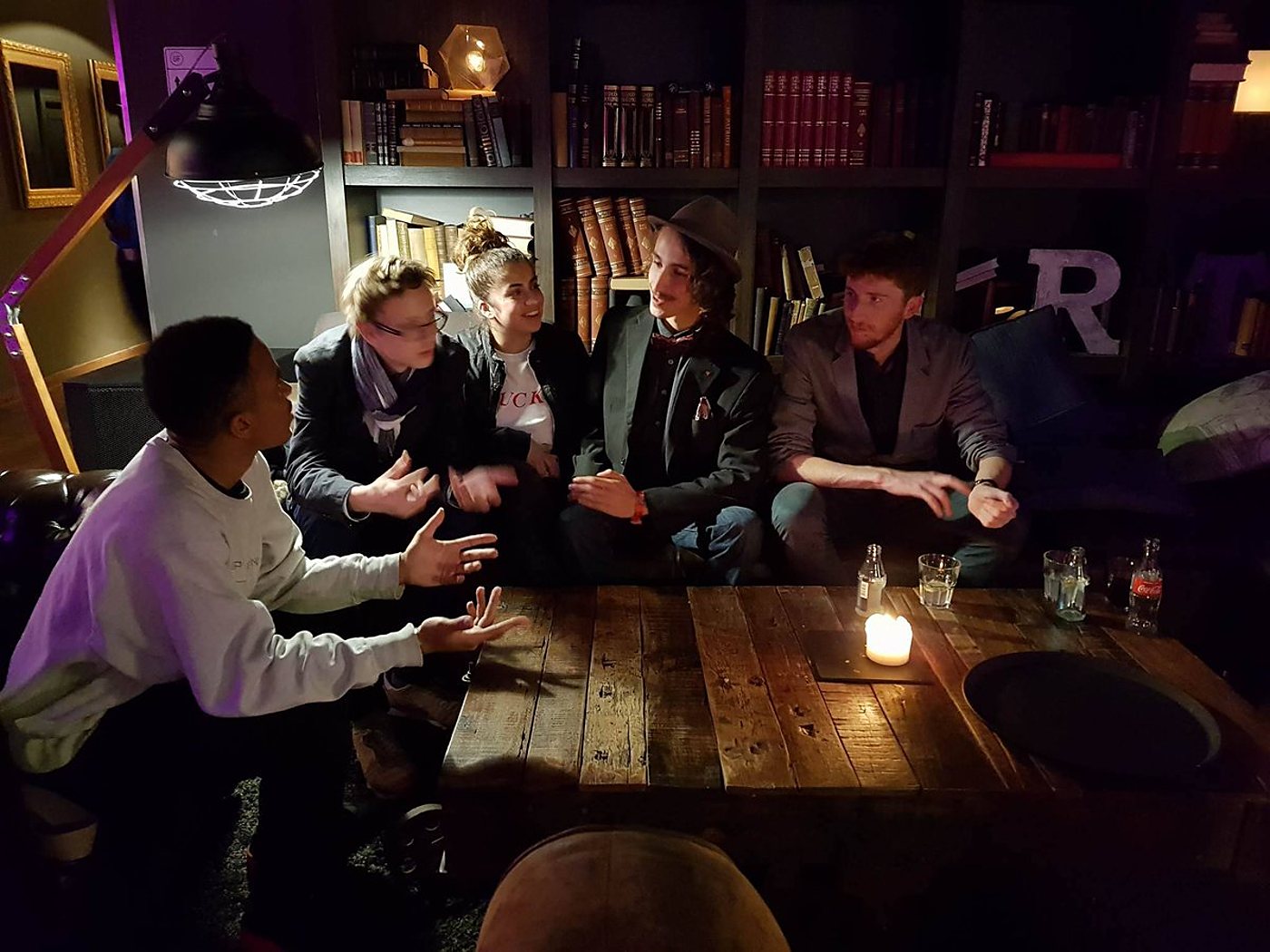 Cinq jeunes gens assis dans un salon sombre discutent en gesticulant autour d'une table basse en bois éclairée par une bougie, devant une bibliothèque remplie de livres.