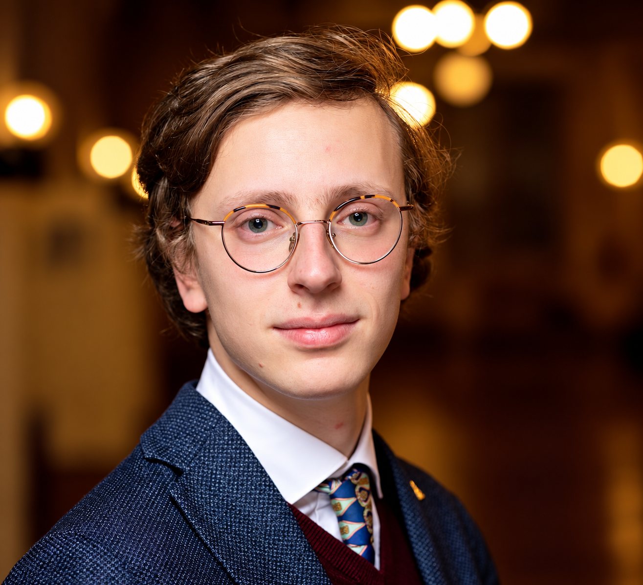 Portrait d'un jeune homme aux cheveux châtains et aux lunettes rondes, vêtu d'une veste bleue et d'une cravate à motifs, devant des lumières floues dorées.