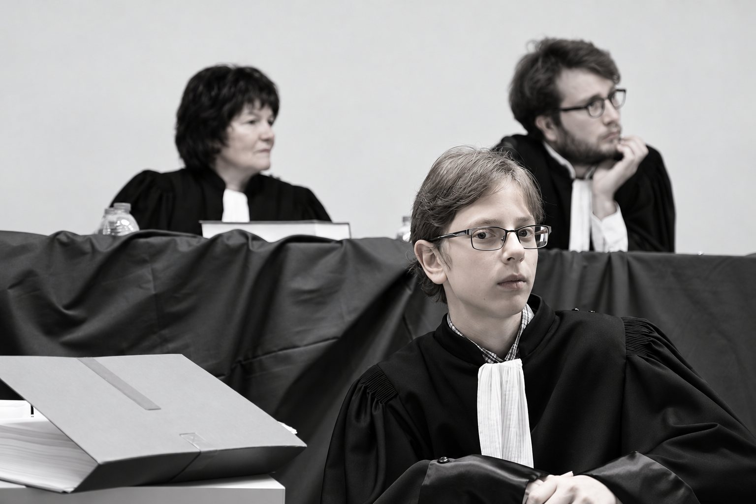 Photo en noir et blanc de trois avocats en robe et rabat blanc, un jeune homme à lunettes au premier plan et deux personnes assises derrière un bureau drapé.
